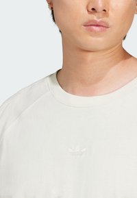 T-shirt en coton beige clair avec un col rond, présentant un logo en relief subtil sur la poitrine et des manches raglan. Texture douce.