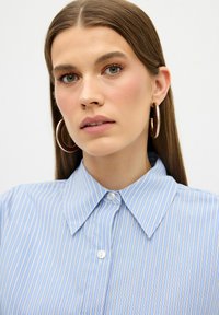 Camicia a righe blu e bianche con bottoni, caratterizzata da un colletto classico e tessuto strutturato. Il modello indossa orecchini a cerchio grandi, con pelle liscia e capelli lisci.