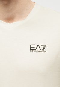 Κοντινή λήψη ενός ανοιχτό μπεζ πουκάμισου με V λαιμόκοψη και μαύρο λογότυπο "EA7 Emporio Armani" στο αριστερό μέρος του στήθους.