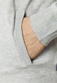 Hand delvis inne i framfickan på en ljusgrå stickad tröja med ribbade ärmslut och fickkanter.