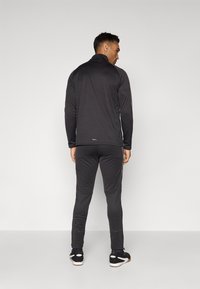 Vīriešu sporta apģērbs melnā krāsā, kas sastāv no zīmola ar rāvējslēdzēju, garām piedurknēm un pieguļošām biksēm. Gluda tekstūra ar diskrētu zīmolu un bez redzamām rakstu telpām.