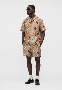 Άνδρας που φοράει μπεζ κοντομάνικο πουκάμισο και σορτς με πολύχρωμο ρομποτικό σχέδιο, λευκές κάλτσες, μαύρα μοκασίνια και σκούρα γυαλιά ηλίου, στέκεται με τα χέρια στις τσέπες.