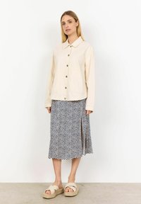 Beige button-up jacket with two front pockets, paired with a knee-length skirt featuring a blue chevron pattern and light-colored heeled sandals.