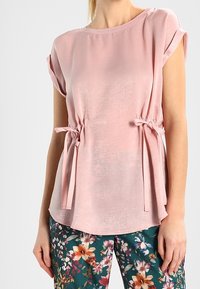 Blusa rosa a maniche corte con scollatura rotonda, lati con cordino e una texture liscia, abbinata a pantaloni con motivo floreale.