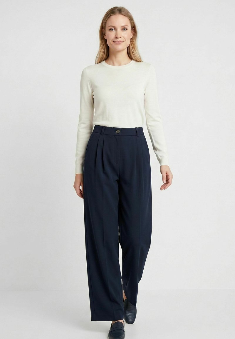 Femme portant un haut blanc à manches longues, un pantalon large taille haute bleu marine et des mocassins foncés, debout devant un fond clair uni.