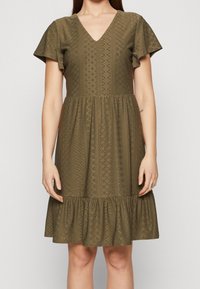 Oliivinvihreä mekko, jossa V-aukko, lyhyet kellohihat, teksturoitu kangas, jossa on timantinmuotoisia kuvioita, ja röyhelöinen helma.