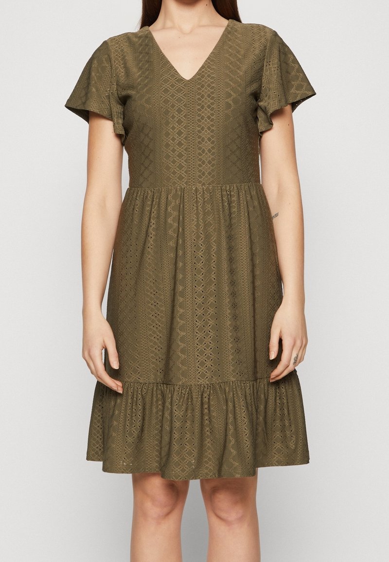 Oliivinvihreä mekko, jossa V-aukko, lyhyet kellohihat, teksturoitu kangas, jossa on timantinmuotoisia kuvioita, ja röyhelöinen helma.