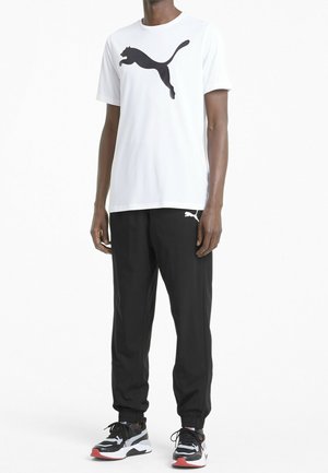 Witte T-shirt met korte mouwen met een zwart Puma-logo, gecombineerd met zwarte joggingsbroek en zwarte sneakers met grijze en rode accenten.