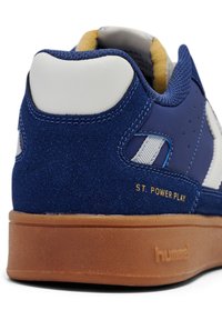 Blå mockaskor med vit läderhäll, tan gummisula och guldtexten "ST. POWER PLAY." Släta tygfoder synliga.