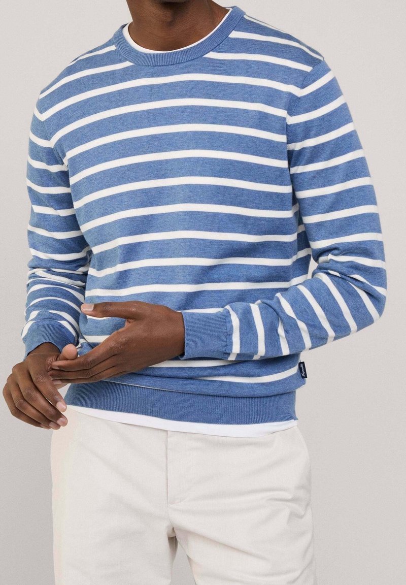 Homme portant un pull à manches longues et col rond avec des rayures horizontales blanches et bleues, associé à un pantalon clair. Mains positionnées en plein geste.