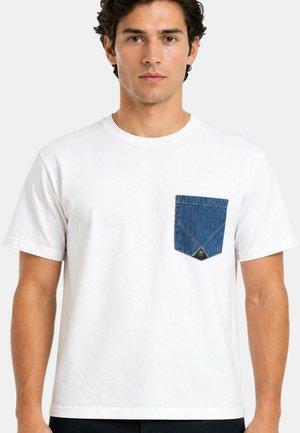 Uomo che indossa una maglietta bianca semplice a maniche corte con una tasca sul petto in denim blu su uno sfondo chiaro uniforme.