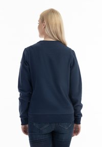 Felpa blu navy con collo rotondo e maniche lunghe, caratterizzata da una texture morbida e polsini a coste. Indossata con jeans scuri, vista da dietro.