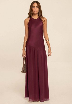 Mujer con cabello largo y ondulado, que lleva un vestido maxi burdeos sin mangas, sosteniendo un bolso marrón con textura, de pie frente a un fondo liso.