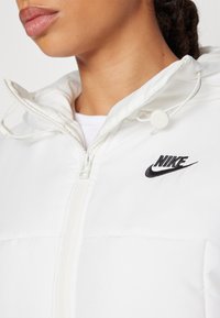 Vit Nike-jacka med dragkedja, huva och broderad logotyp. Mjuk, slät textur med en strukturerad passform och justerbara dragkedjor.