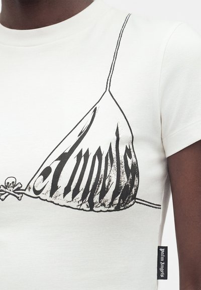 Κοντινή λήψη λευκού T-shirt με μαύρο τυπωμένο σχέδιο που μοιάζει με πάνω μέρος μπικίνι και κρανίο με σταυρωμένα κόκαλα, που φοριέται από άτομο με σκουρόχρωμο δέρμα.