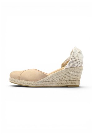 Alpargata beige con cuña, puntera cerrada y correa trenzada en el tobillo sobre fondo blanco.
