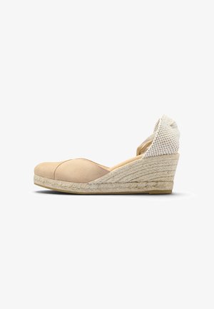 Alpargata beige con cuña, puntera cerrada y correa trenzada en el tobillo sobre fondo blanco.