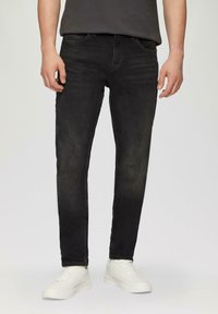 Jean en denim noir à coupe slim, avec un design à cinq poches et des détails légèrement décolorés. Porté avec des baskets blanches et un t-shirt gris.