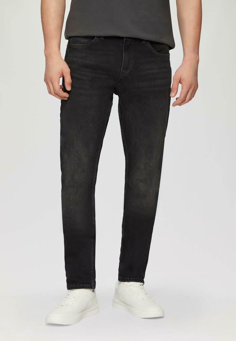 Jean en denim noir à coupe slim, avec un design à cinq poches et des détails légèrement décolorés. Porté avec des baskets blanches et un t-shirt gris.