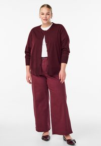 Weinroter Cardigan mit Knopfleiste, kombiniert mit passenden weit geschnittenen Hosen. Der Stoff wirkt weich mit einer glatten Textur. Schwarze Ballerinas runden das Outfit ab.