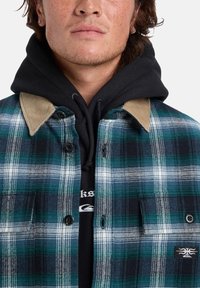 Jonge man in een groen-blauwe geruite jas met een lichtbruine corduroy kraag over een zwarte hoodie met witte tekst.