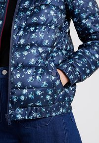 Marinebloemige pufferjack met polyester materiaal, voorzien van lichtblauwe bloemen, gestikt textuur en elastische manchetten. Ritsluiting en zakken.