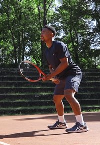 Τennis παίκτης με ναυτικό μπλε μπλουζάκι και σορτς, κρατώντας μια κόκκινη και λευκή ρακέτα. Φοράει γκρι παπούτσια τένις με λευκές λεπτομέρειες σε χωμάτινη επιφάνεια.