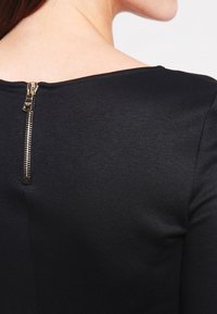 Top negro con una textura suave, escote amplio y una cremallera trasera visible con herrajes metálicos.