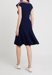Mujer vistiendo un vestido de longitud hasta la rodilla en color azul marino con rayas rojas y blancas en los hombros, combinado con zapatillas blancas, de pie frente a un fondo liso.
