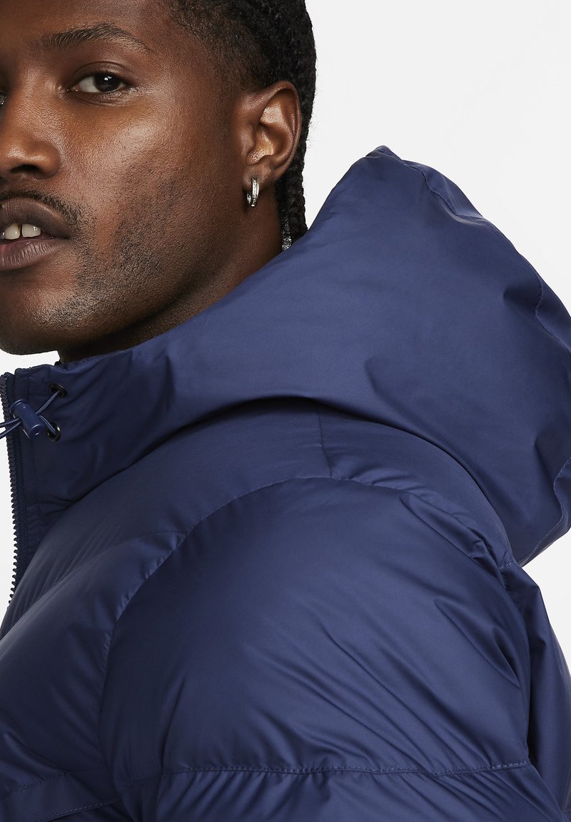 Marineblaue Daunenjacke mit Kapuze, die eine glatte, glänzende Textur, horizontale Nähte und einen Reißverschluss an der Vorderseite aufweist.