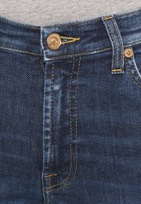 Jeans de denim azul oscuro con un botón de cobre y costuras amarillas. Diseño estructurado con corte recto y textura sutil.