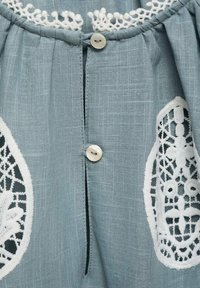 Blusa di lino azzurro chiaro con dettagli in pizzo bianco sui lati, bottoni decorativi sul davanti e tessuto leggermente strutturato.