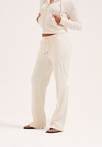 Γυναίκα με κρεμ φούτερ με φερμουάρ και παντελόνι φαρδύ φόρμας, φορόντας μαύρα σανδάλια με τακούνι, στέκεται με τα χέρια στην τσέπη.