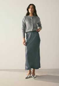 Grau gestrickte Strickjacke mit rundem Ausschnitt und Knöpfen, kombiniert mit einem langen, fließenden tealfarbenen Unterrock. Silberne Absatzschuhe vervollständigen das Outfit.