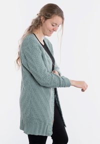 Dzianinowy kardigan w jasnym odcieniu turkusowym, z teksturowanym wzorem w jodełkę, z otwartym przodem i ściągaczami na rękawach, noszony na czarnej bluzce i ciemnych spodniach.
