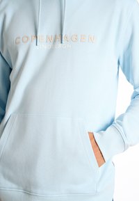 Sudadera azul claro con un bolsillo frontal, que presenta la impresión en blanco de "COPENHAGUE" y "LINDBERGH". Fabricada con un tejido suave y un corte relajado.