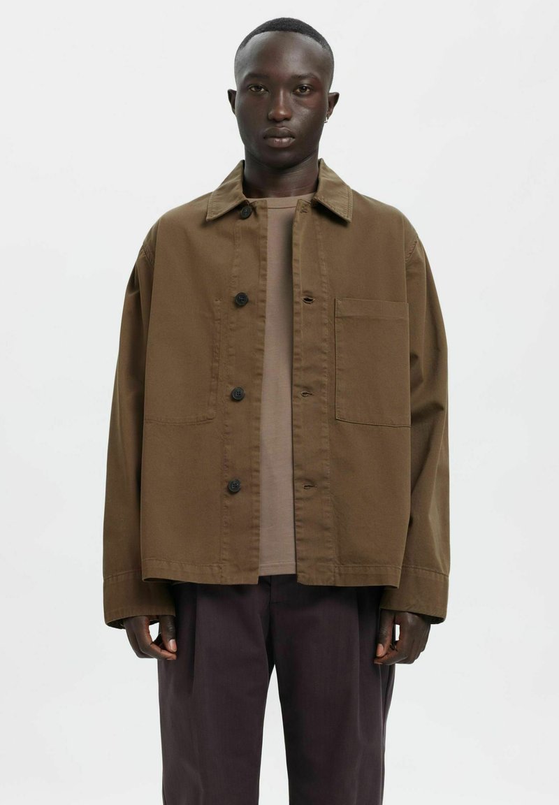 Brązowa bawełniana kurtka z zapięciem na guziki, kołnierzem i jedną kieszenią na piersi, noszona na beżowej koszuli. Czarnie spodnie dopełniają strój.