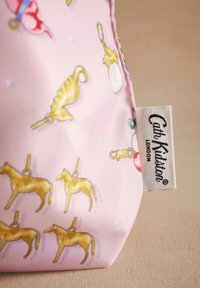 Ružový textilný vrecúško s jazdeckými zlatými vzormi a rôznymi ikonami. Štítok hovorí "Cath Kidston London." Hladká textúra, hravý vzor.