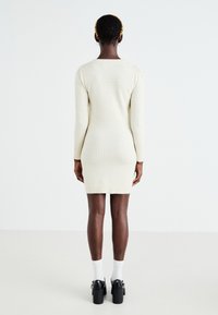Robe en tricot crème à manches longues, silhouette ajustée, motif texturé et décolleté rond, assortie à des chaussures noires à talons hauts et des chaussettes blanches.