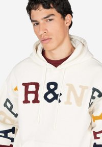 Sudadera crema con letras de aplique multicolores en varias tipografías, con capucha ajustable y un acabado suave y texturizado.