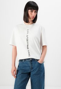 Λευκό βαμβακερό T-shirt με κάθετη εκτύπωση μαύρου λογότυπου που γράφει "KARL LAGERFELD JEANS". Συνοδεύεται από μπλε τζιν σε χαλαρή γραμμή.