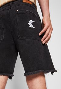 Mustat farkkushortsit, joissa on repaleinen helma, kaksi takataskua ja kirjailtu valkoinen logo. Materiaali vaikuttaa tukevilta ja siinä on matta pinta.