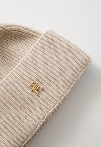 Gorro de punto beige con una textura acanalada y un detalle de emblema dorado. La forma es redondeada, enfatizando un diseño y material elegantes.