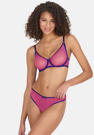 Frau mit lockigem Haar, die einen rosa und marineblauen durchsichtigen BH und passende Slips trägt und mit einer Hand in der Hüfte vor weißem Hintergrund posiert.