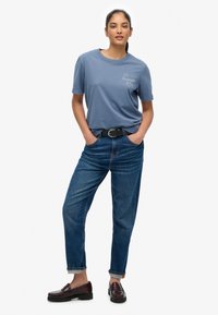Blå bomulds-T-shirt med korte ærmer og rund hals; har en hvid logo. Blå løstsiddende denimjeans med bælte. Brune loafers.