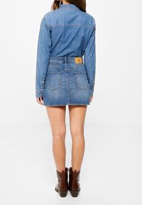 Abito camicia in denim, blu chiaro, maniche lunghe, vita aderente, due tasche posteriori, etichetta sulla vita, lunghezza sopra il ginocchio, abbinato a stivaletti marroni.