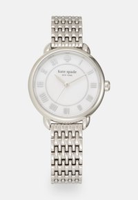Silverfärgad klocka med ett band av metallnät. Rund vit urtavla har romerska siffror. Märkesnamnet "kate spade" visas under 12-timmarsmarkeringen.