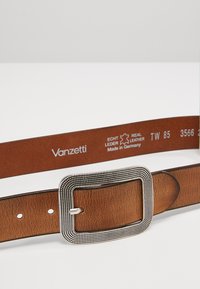 Ceinture en cuir marron avec une boucle en métal argenté texturée. Elle présente une surface lisse, plusieurs trous et une étiquette de marque à l'intérieur.