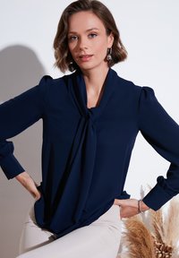 Marineblauw blouse met een geknoopte halslijn, lange mouwen met geplooide schouders en een gladde textuur. Gecombineerd met lichte broek.