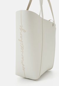 Sac fourre-tout en simili cuir blanc texturé avec une finition lisse, arborant un logo cursif subtil en beige clair sur le côté et doté de deux poignées.